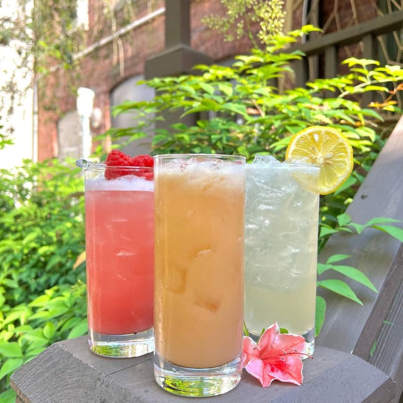 Three colorful cocktails, garnished with raspberries and lemon, sit on a wooden ledge outdoors.