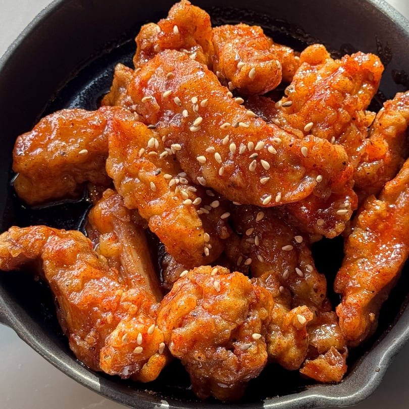 Close-up of Korean fried chicken pieces coated in a glossy red sauce and sesame seeds in a black bowl.
