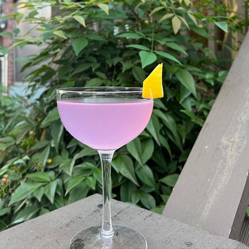 A vibrant purple cocktail in a coupe glass, garnished with a lemon wedge, set against lush greenery.